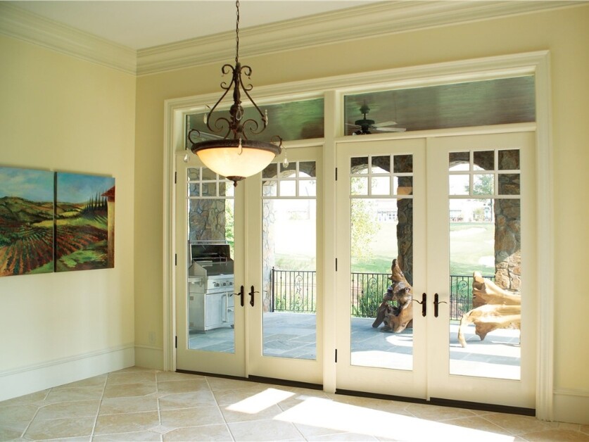 White French doors with transom windows and decorative handles leading to a covered patio with outdoor kitchen. Interior features beige tile flooring, cream walls, and vineyard artwork.